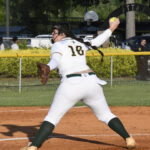 A softball pitcher in a white uniform prepares to throw the ball during a game, captured from behind on a sunny outdoor field.