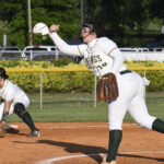 A softball pitcher in a white and green uniform throws a pitch while a teammate prepares to field on a dirt field.
