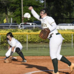 A softball pitcher throws the ball while a teammate in the infield prepares to catch. Both wear white uniforms with green socks. Outdoor field setting.