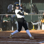 A softball player in black and white uniform swings a bat during a game, with a chain-link fence and spectators in the background.
