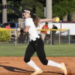 A softball player in uniform throws a yellow softball during a game on a dirt field, with spectators and trees in the background.