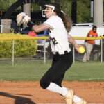 A softball player in uniform is pitching on a dirt field, wearing a face mask, with spectators and vehicles in the background.