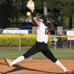 A softball pitcher in a white and black uniform throws a pitch mid-air on a dirt field, with trees and cars in the background.