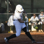 Softball player in white uniform swings bat at a pitch during a game, with teammates watching from the bench behind a chain-link fence.