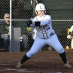 A softball player in white uniform prepares to bunt, holding the bat forward, with spectators and a chain-link fence in the background.