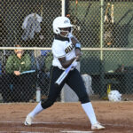 A softball player in uniform swings a bat during a game, standing on a dirt field with a chain-link fence and spectators in the background.