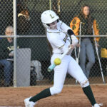 A softball player in white uniform swings at a pitched ball, with spectators and a chain-link fence in the background.