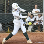 A softball player in white uniform swinging a bat at a pitch, with teammates watching from the dugout behind a chain-link fence.