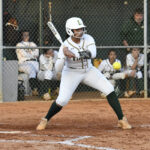 A softball player in uniform prepares to bat, standing at home plate with a bat, while teammates watch from the dugout in the background.