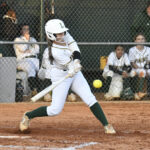A softball player in white uniform swings at a yellow ball during a game, with teammates watching from the dugout in the background.