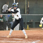 A softball player wearing a helmet prepares to bat at home plate on a dirt field, with a chain-link fence and dugout in the background.