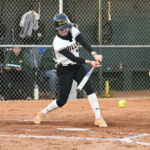A softball player in uniform swings a bat to hit an incoming yellow softball on a dirt field, with a chain-link fence and spectators in the background.