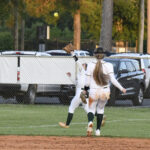 Two softball players in white uniforms chase a fly ball in the outfield, with cars and trees visible in the background.