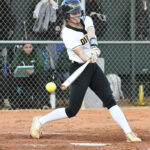 A softball player in uniform hits a pitched ball with a bat during a game, with spectators and a fence visible in the background.