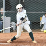 A softball player in white and green uniform swings a bat during a game, with a chain-link fence and spectators in the background.