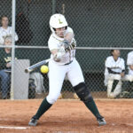 A softball player in uniform swings a bat at an incoming pitch on a dirt field, with teammates and a chain-link fence in the background.