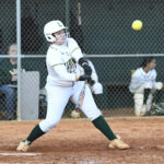 A softball player in uniform swings at a pitch during a game, with teammates watching from the dugout in the background.