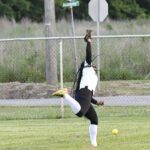 A softball player in black and white uniform extends arm to catch a ball on a grassy field, with a fence and road in the background.
