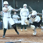 A softball player runs to home plate as the catcher attempts to tag her out, with teammates watching in the background.