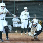 A softball player swings at a pitch while the catcher prepares to catch the ball and teammates watch from the dugout area.