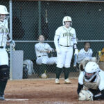A girls’ softball game with players in white uniforms; one batter stands ready, a catcher crouches behind home plate, and teammates watch from the dugout.