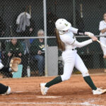 A softball player in white swings at a pitch while the catcher and spectators watch from behind the fence on a dirt field.