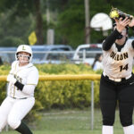 A softball player catches a ball with a glove while another player in white runs toward a base on a grassy field.