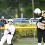 Two female softball players on the field, one preparing to catch a ball and the other running, with spectators and parked cars in the background.