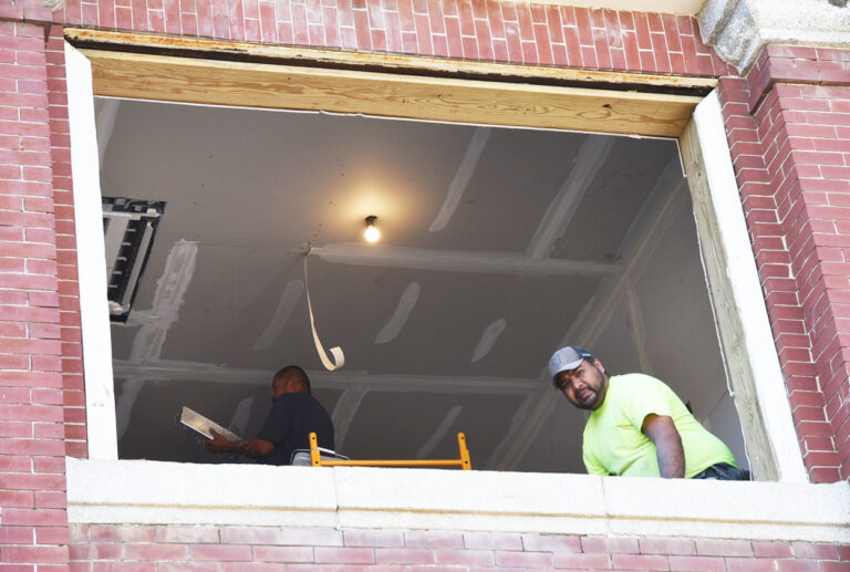 Two construction workers inside a building, one wearing a cap and neon shirt, looking out from a large unfinished window with exposed drywall and a ceiling light.