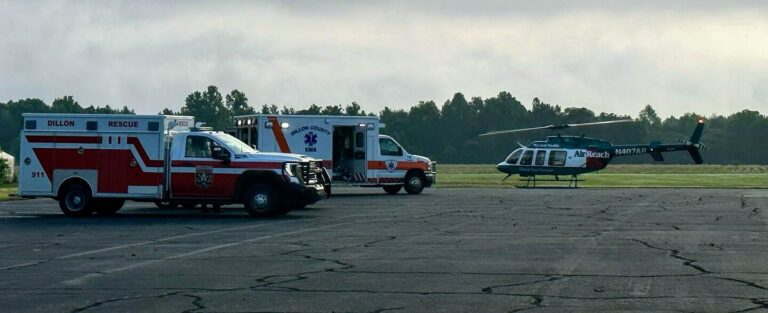 Ambulances and a medical helicopter are parked on an airstrip, prepared for emergency response, with trees in the background under a cloudy sky.