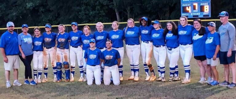 A girls’ softball team in blue uniforms poses together on a grassy field, with some players standing and others kneeling in front, trees visible in the background.