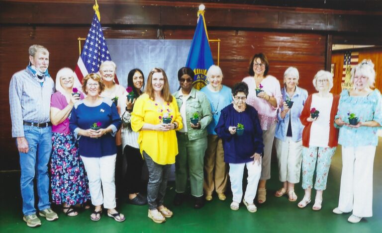 Group of women standing indoors, holding small plants, with American and blue flags in the background.