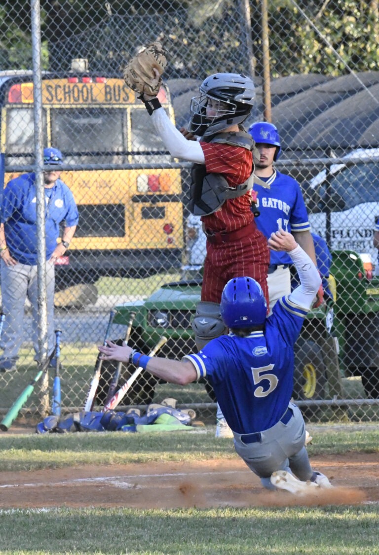 Baseball catcher in red gear jumps to catch the ball as a player in blue uniform slides toward home plate, with coaches and equipment visible in the background.
