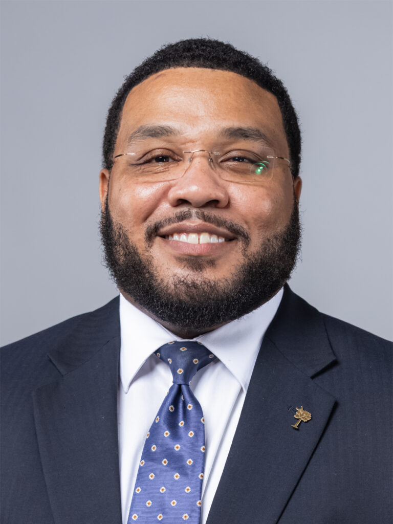 Smiling man with short hair, beard, and glasses wearing a dark suit, white shirt, and blue polka-dot tie, posed against a light gray background.