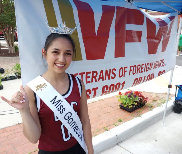 Young woman wearing a crown and "Miss Cotter 2015" sash, smiling in front of a VFW banner at an outdoor event.