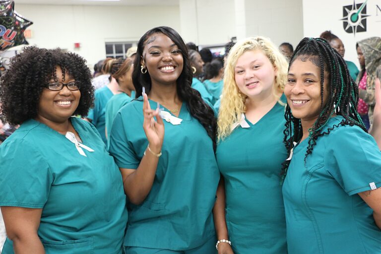 Four women in green scrubs smiling and posing together indoors, possibly at a medical or nursing school event.