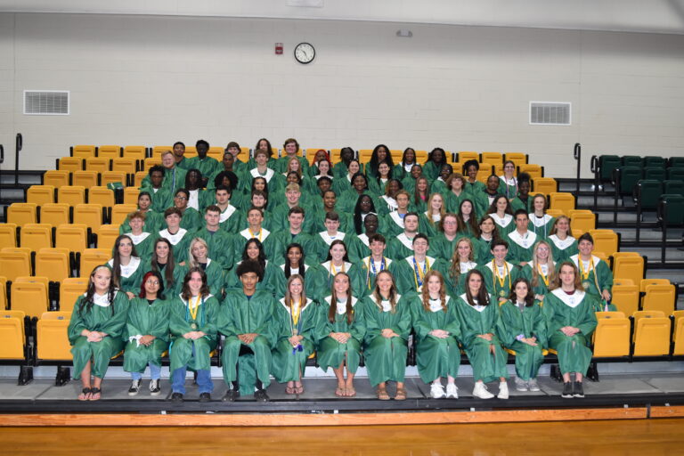 A large group of graduates in green gowns pose together on bleachers inside a gymnasium for a class photo.