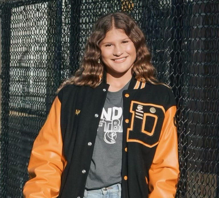 Smiling young woman with wavy hair wears a black and orange varsity jacket, standing outdoors in front of a chain-link fence.