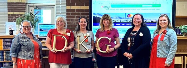 Four women stand indoors, smiling and holding large gold letters "D," "K," and "G," with a presentation screen in the background.