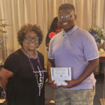 older woman and a young man stand indoors; the young man holds a certificate and both are smiling at the camera.