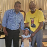 Two men and a young girl pose together indoors; one man wears a yellow shirt with a printed photo, the girl holds a small brown bag.