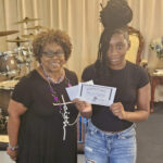Two women standing indoors, one holding a certificate and envelope, both smiling at the camera. Musical instruments and a chair are visible in the background.