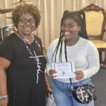 Two women smiling indoors, one holding a certificate. Both are casually dressed, with visible crosses on a necklace and a handbag. They appear to be at an event or ceremony.