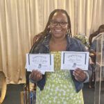 Smiling woman wearing glasses and a green dress holds two certificates, standing indoors with a beige curtain in the background.