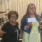 Two smiling women stand indoors; one holds a piece of paper while the other wears glasses and a necklace. Drums and beige curtains are visible in the background.