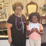 older woman stands next to a smiling child holding a certificate, both posing indoors with musical instruments and elegant chairs in the background.