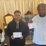 Three people standing indoors, holding certificates, with a woman sitting behind them and cream-colored curtains in the background.