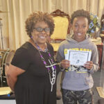 A woman and a young person stand indoors, smiling at the camera. The young person is holding a certificate. Musical instruments and chairs are visible in the background.