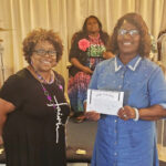 Two women smiling, one holding a certificate, posing together indoors with another seated woman in the background.