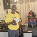A man in a yellow "STAFF" t-shirt holding a card stands in a decorated room, while two women sit in the background.